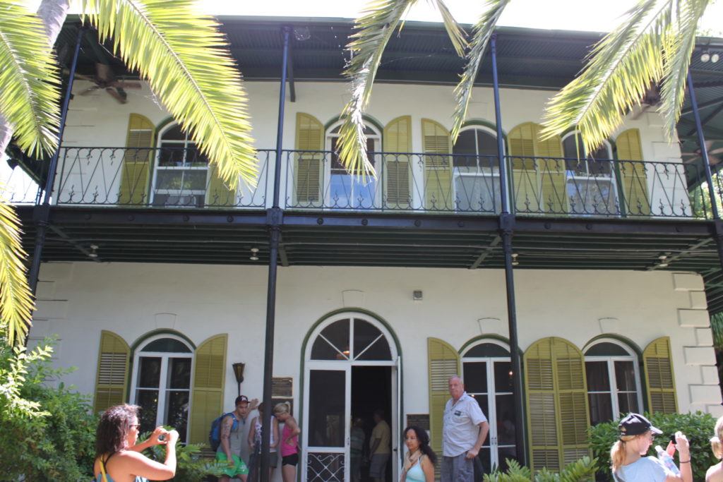 A white stone building with a metal upper porch and green shutters flanked by palm trees at  Ernest Hemingway Home & Museum in Key West