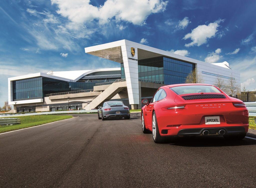 Two red and silver sports cars drive on the track at the modern white building at the Porsche Experience Center